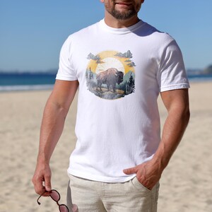 May include: White t-shirt with a vintage-style graphic of a buffalo in a forest setting with a sun and clouds. The graphic is in shades of yellow, orange, brown, and green.