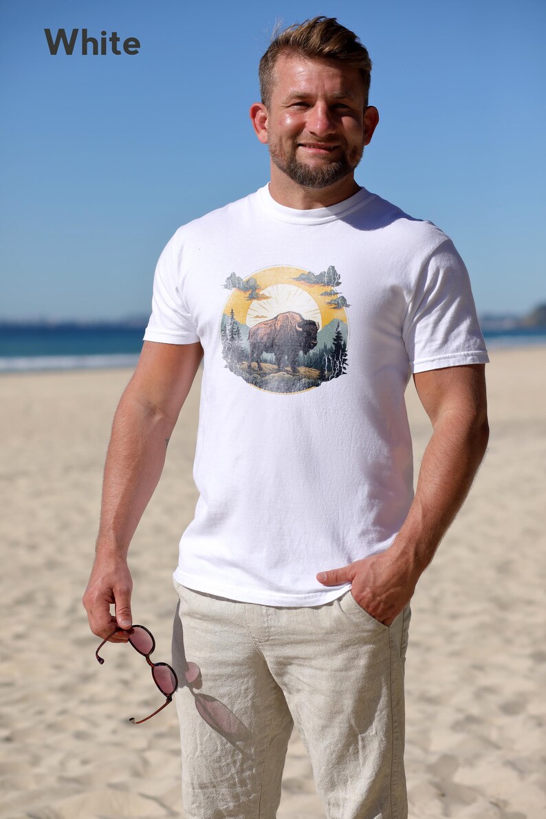 May include: White t-shirt with a vintage-style graphic of a buffalo in a forest setting with a sun and clouds. The graphic is in shades of yellow, orange, brown, and green.
