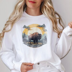 May include: White long-sleeve sweatshirt with a vintage-style graphic of a brown bison standing in a forest with a yellow sun and clouds in the background.