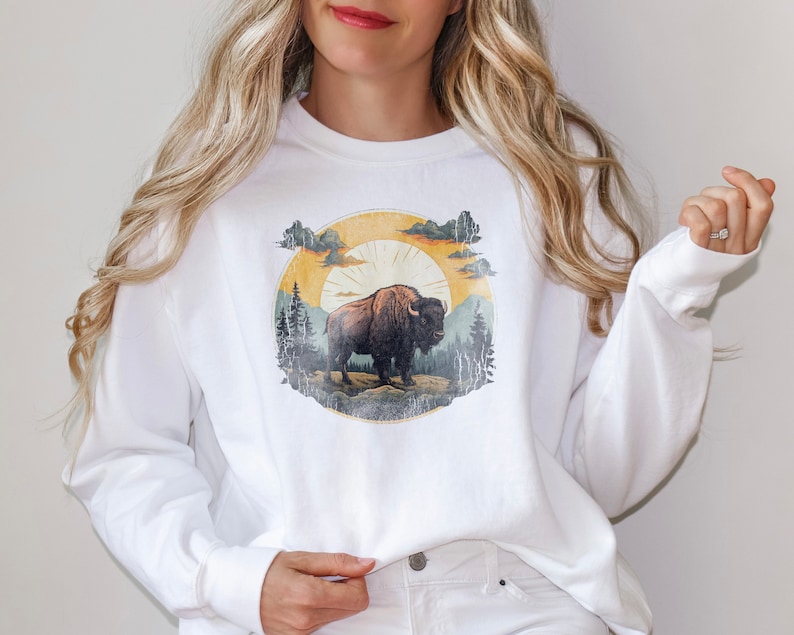 May include: White long-sleeve sweatshirt with a vintage-style graphic of a brown bison standing in a forest with a yellow sun and clouds in the background.