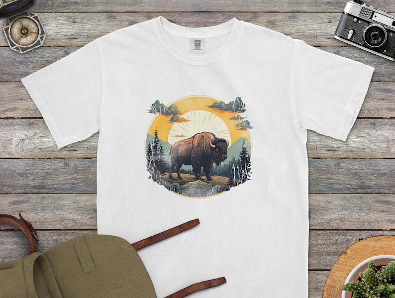 This white t-shirt features a striking vintage-inspired bison design, set against a warm-toned sunset with scenic mountain and forest details. The artwork exudes a rugged, adventurous aesthetic, perfect for nature lovers, outdoor enthusiasts, and those who appreciate wildlife-inspired fashion. Styled alongside a canvas backpack, vintage camera, and rustic wood textures, this setup captures the essence of exploration and wanderlust.
