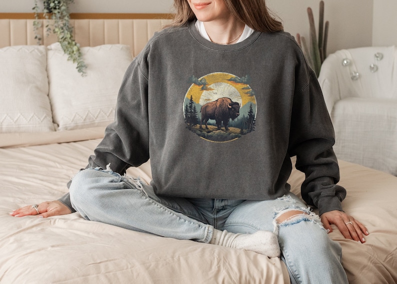A woman wearing a dark gray oversized sweatshirt featuring a detailed, vintage-style illustration of a bison standing on rugged terrain, surrounded by pine trees. The design is set within a circular frame, depicting a warm, golden sunset sky with clouds. She pairs the sweatshirt with light-wash, distressed jeans and cozy socks while sitting cross-legged on a neutral-toned bed. The soft, bohemian-inspired room setting enhances the relaxed and nature-inspired aesthetic of the outfit.