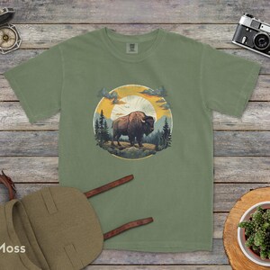 May include: A green t-shirt with a graphic of a brown bison standing in a forest with a sunset in the background. The graphic is surrounded by a circle with a yellow sun and a blue sky.