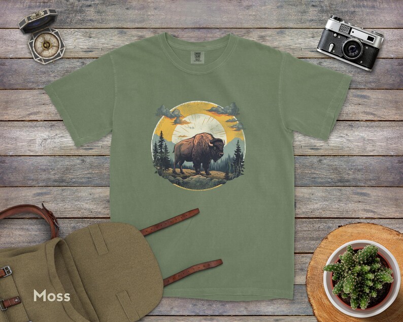 May include: A green t-shirt with a graphic of a brown bison standing in a forest with a sunset in the background. The graphic is surrounded by a circle with a yellow sun and a blue sky.
