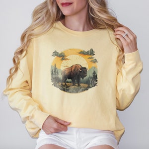 May include: A yellow sweatshirt with a vintage-style graphic of a brown bison standing in a forest with a sun and clouds in the background.
