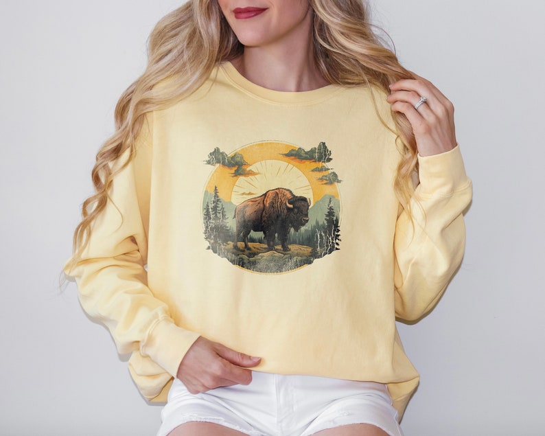 May include: A yellow sweatshirt with a vintage-style graphic of a brown bison standing in a forest with a sun and clouds in the background.