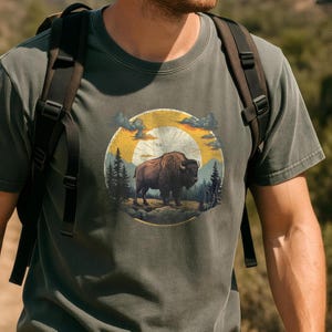 May include: A dark green t-shirt with a vintage-style graphic of a brown buffalo standing in a forest scene with a yellow sun and clouds.