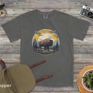 May include: A dark gray t-shirt with a vintage-style illustration of a bison standing in a forest with a sunset in the background.