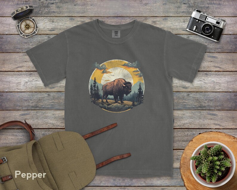 May include: A dark gray t-shirt with a vintage-style illustration of a bison standing in a forest with a sunset in the background.