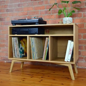 May include: A light oak record player stand with a turntable, CD player, and vinyl record storage. The stand has angled legs and a mid-century modern design. Two books are visible on the right side. A plant in a white pot sits on top.