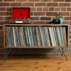 May include: A wooden record player stand with a red vintage record player on top and a shelf full of vinyl records. The stand has hairpin legs and is sitting on a wooden floor.