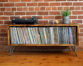 May include: A wooden record player stand with a black turntable on top and a shelf below for storing vinyl records. The stand has hairpin legs and is in front of a brick wall.