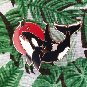 May include: Enamel pin depicting a black and white orca whale with a red sun. The pin has gold-coloured outlines and is set against a green and white tropical leaf pattern.