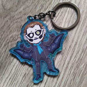 May include: A cartoon keychain depicting a character in a dark suit with bat-like wings. The character has a white face with black accents and a green eye. The keychain has a silver keyring and a black chain.