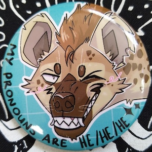May include: A circular badge featuring a cartoon hyena with brown and tan fur, a wide grin, and the text "MY PRONOUNS ARE HE/HE/HE" on a teal background. The badge is set against a black and white patterned fabric.