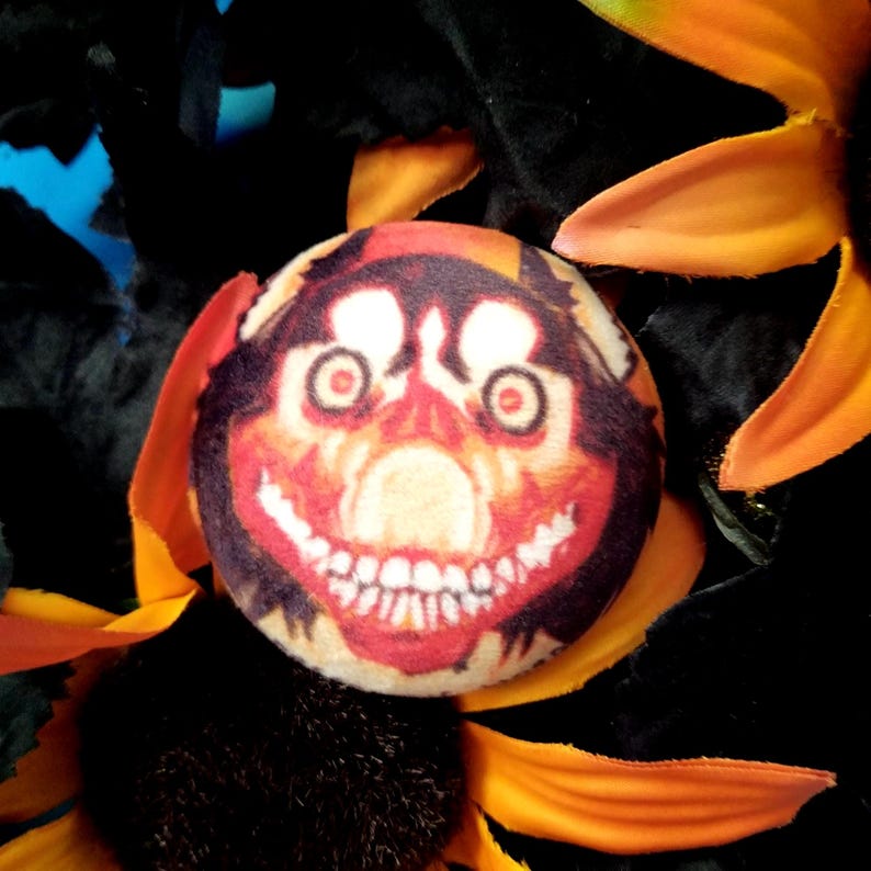 May include: A round button with a creepy face design. The button features a smiling face with red eyes, a wide grin, and a black and orange background. The button is surrounded by orange and yellow flower petals.