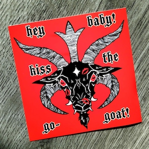 May include: A square, red sticker with a black and white illustration of a goat's head. The text "hey baby! kiss the goat!" is written around the goat's head in a gothic font. The goat has red eyes and a star on its forehead. A dark, gothic design.