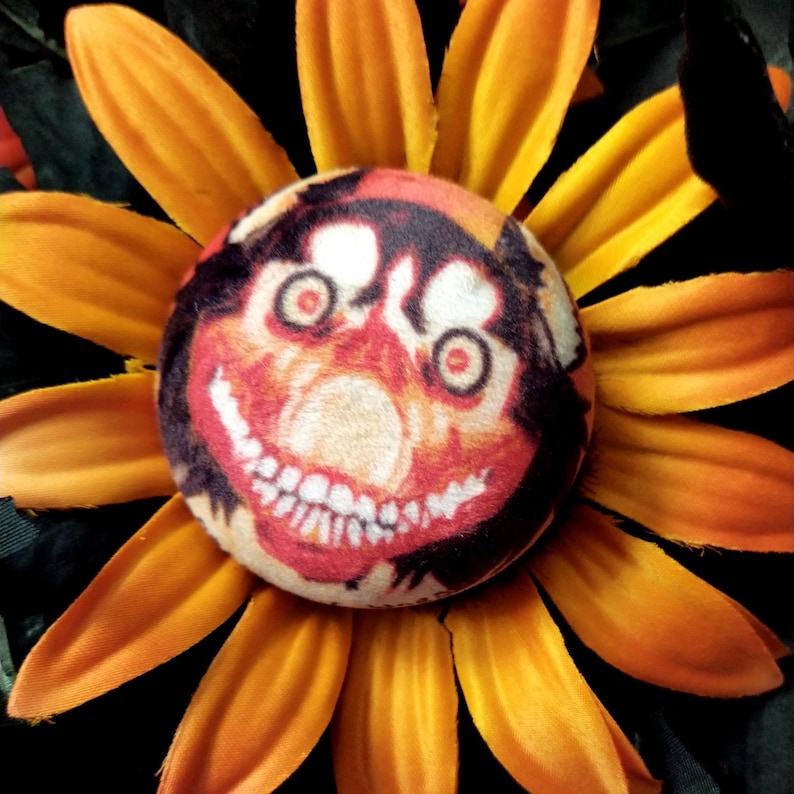 May include: A unique, handmade sunflower with orange petals and a central design featuring a smiling face with red, white, and black details. The flower has a whimsical, slightly unsettling appearance.