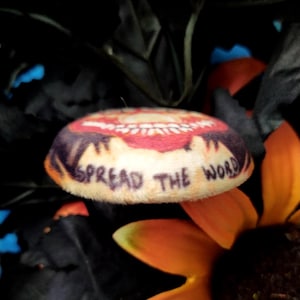 May include: A round, plush item with a cream-colored base and a red and brown design on top. The words "SPREAD THE WORD" are printed on the side. The item is set against a backdrop of dark leaves and an orange flower.