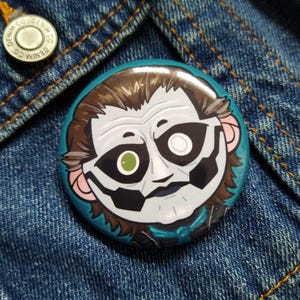 May include: A round button with a cartoon illustration of a character with white face paint, brown hair, and a teal background. The button is set against a blue denim background.
