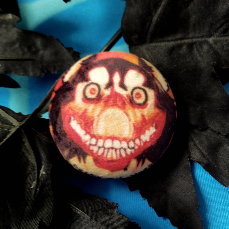 May include: A round, plush toy with a smiling face design. The face features red eyes, a wide grin with visible teeth, and a color palette of orange, red, and brown. The toy is set against a blue background with black decorative elements.