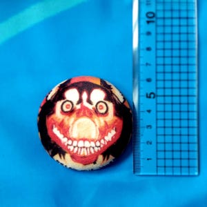 May include: A round button with a creepy face design. The button features a red, orange, and black color scheme with a large, toothy grin. A ruler is next to the button, showing measurements in inches.