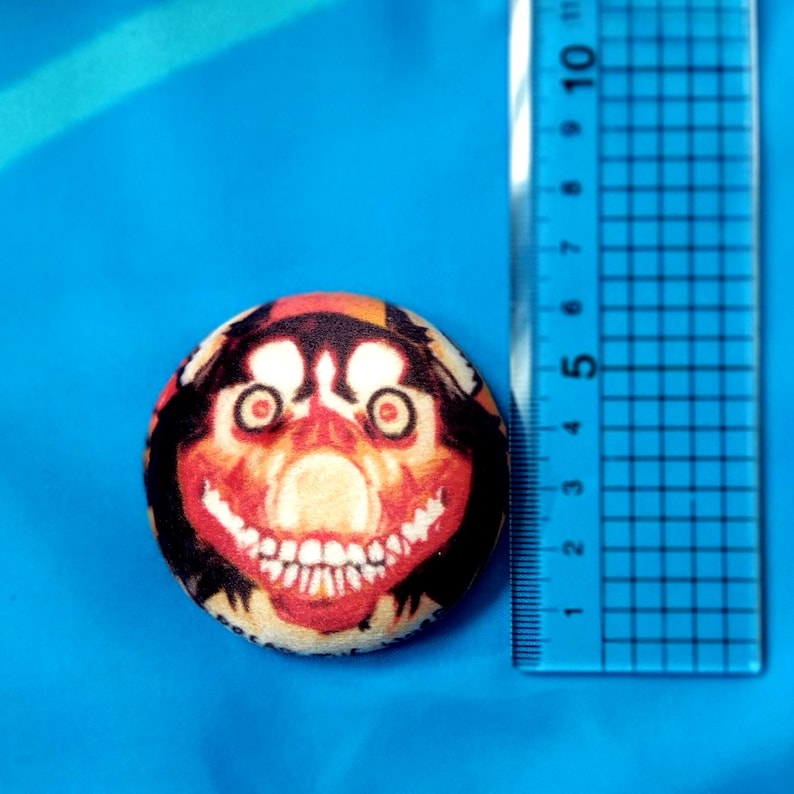 May include: A round button with a creepy face design. The button features a red, orange, and black color scheme with a large, toothy grin. A ruler is next to the button, showing measurements in inches.