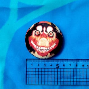 May include: A round button with a creepy face design. The button features a red, black, and beige color scheme, with a large, unsettling grin and wide eyes. The button is approximately 2 inches in diameter.