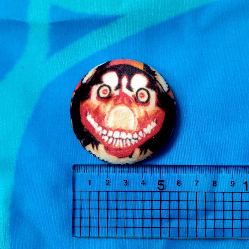 May include: A round button with a creepy face design. The button features a red, black, and beige color scheme, with a large, unsettling grin and wide eyes. The button is approximately 2 inches in diameter.