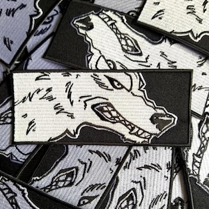 May include: Embroidered patches with a snarling wolf head design. The patches have a black border and a white wolf head with black detailing. The patches are rectangular and appear to be made of fabric. The patches are scattered across a surface.