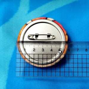 May include: A round button pin with a safety pin back, measuring approximately 2 inches in diameter. The button has a colorful, patterned edge and a silver metal back. The pin is placed on a blue surface.