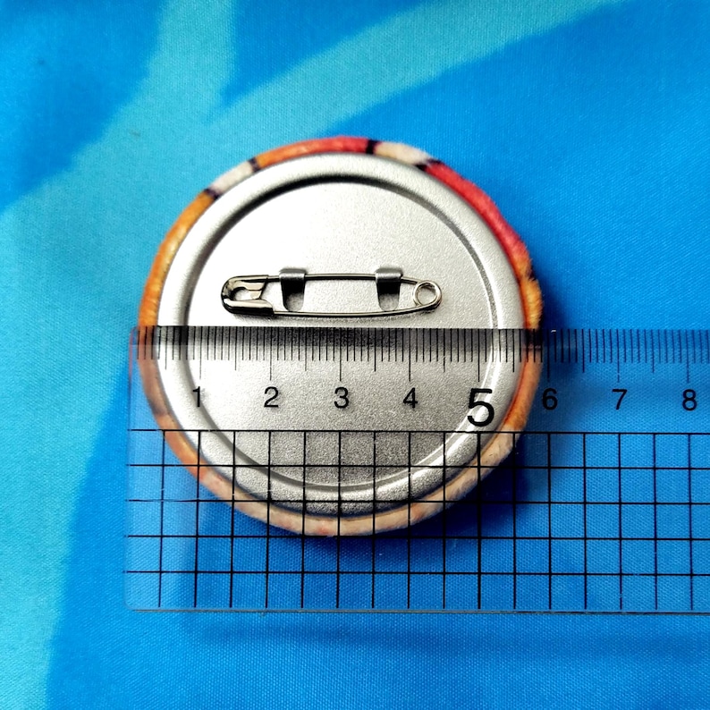 May include: A round button pin with a safety pin back, measuring approximately 2 inches in diameter. The button has a colorful, patterned edge and a silver metal back. The pin is placed on a blue surface.