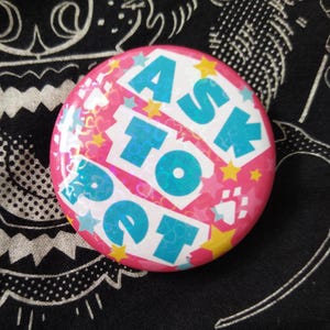 May include: A round, pink button with the text "ASK TO PET" in large, turquoise letters. The button has a holographic effect and is decorated with stars and paw prints.