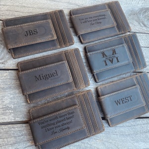 May include: Several gray leather money clips with personalized engravings. One clip reads "JBS", another "Miguel", and others feature names or phrases like "You are my everything..." and "WEST". The clips are arranged on a wooden surface.