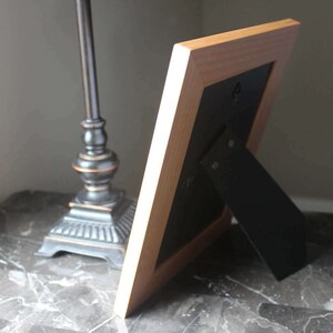 May include: A light-coloured wooden picture frame with a black backing and a black easel stand. The frame is leaning against a dark surface, with a decorative lamp in the background.