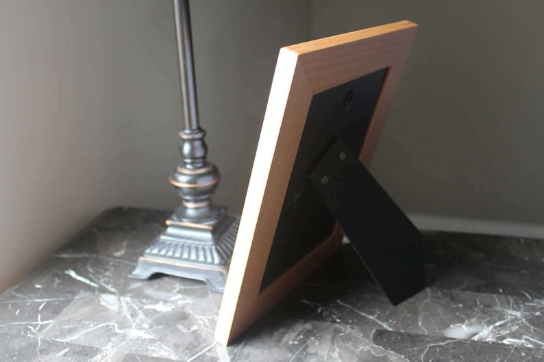 May include: A light-coloured wooden picture frame with a black backing and a black easel stand. The frame is leaning against a dark surface, with a decorative lamp in the background.