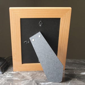 May include: A light-coloured wooden picture frame with a black backing. The frame has a built-in easel for standing. The back of the frame has two metal hangers for wall mounting.