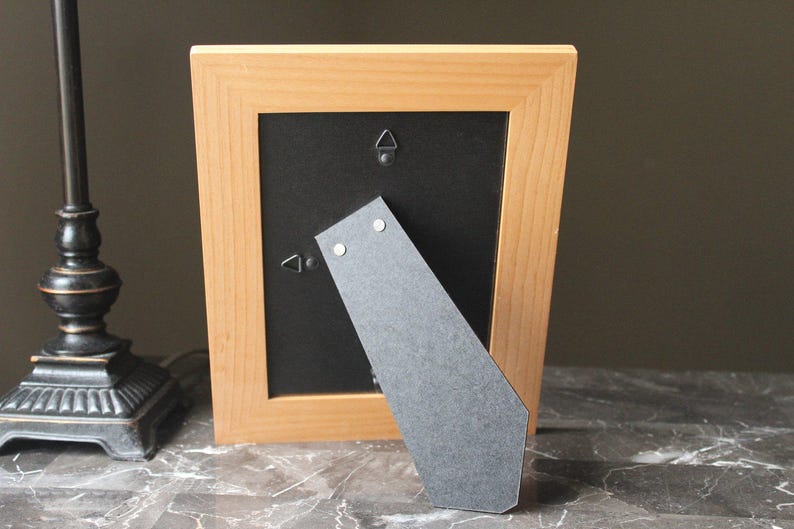 May include: A light-coloured wooden picture frame with a black backing. The frame has a built-in easel for standing. The back of the frame has two metal hangers for wall mounting.