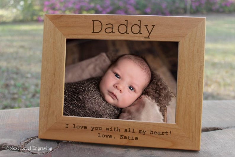 May include: A light wooden picture frame with the word "Daddy" engraved at the top. The frame contains a photo of a baby in a brown blanket. The text "I love you with all my heart! Love, Katie" is engraved at the bottom.