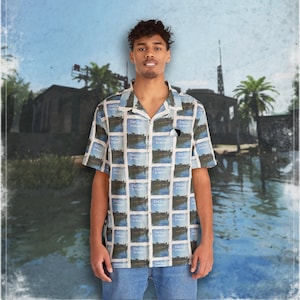 May include: A man is wearing a light blue and white short-sleeved button-down shirt with a repeating pattern of a landscape scene. The shirt has a collar and a button-up front. The man is standing in front of a blurry background of a tropical scene.