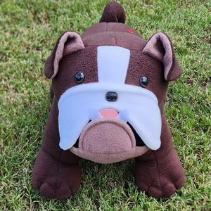 May include: A plush bulldog toy made of dark brown fleece fabric with a white patch extending from the forehead to the muzzle. It features black plastic eyes, a small black nose, a pink tongue, and two small white teeth protruding from the mouth.