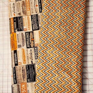 May include: A folded fabric roll featuring two distinct patterns. One side displays vintage apothecary labels with "POISON" warnings. The other side showcases a repeating orange, black, and cream chevron design. The fabric is designed to have a distressed appearance.