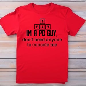 May include: A red t-shirt with black text and keyboard arrow graphics. The text reads "IM A PC GUY, don't need anyone to console me". The shirt is displayed on a light wood grain surface.