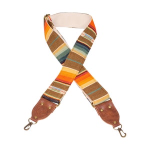 May include: A colorful camera strap with a brown leather adjustable strap and metal clasps. The strap is made of woven fabric with orange, yellow, blue, and brown stripes.
