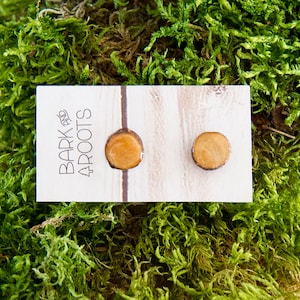 May include: A pair of small, round wooden earrings with a natural wood grain pattern. The earrings are a light brown color and have a smooth, polished finish. The earrings are displayed on a white card with the text "Bark and Roots" printed on it.