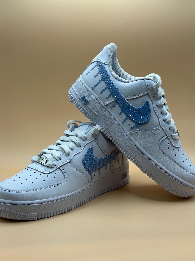 May include: White leather low-top sneakers with a blue rhinestone swoosh and drip design.