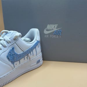 May include: White leather Nike Air Force 1 sneakers with a blue rhinestone swoosh and drip design. The shoes are in a box with the Nike logo and "Air Force 1" text.