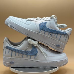 May include: White leather sneakers with a blue rhinestone Nike swoosh and drip design.