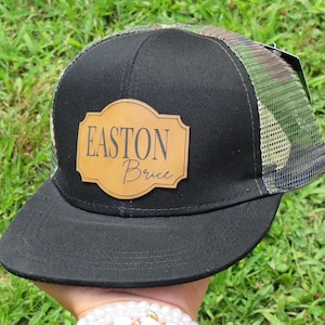May include: Black trucker cap with a camouflage mesh back and a curved peak. A brown leather patch on the front displays the name "Easton Brice". The hat is set against a backdrop of green grass.