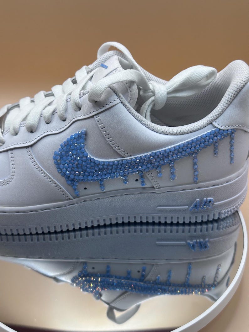 May include: White leather low-top sneakers with a blue rhinestone swoosh and drip design.